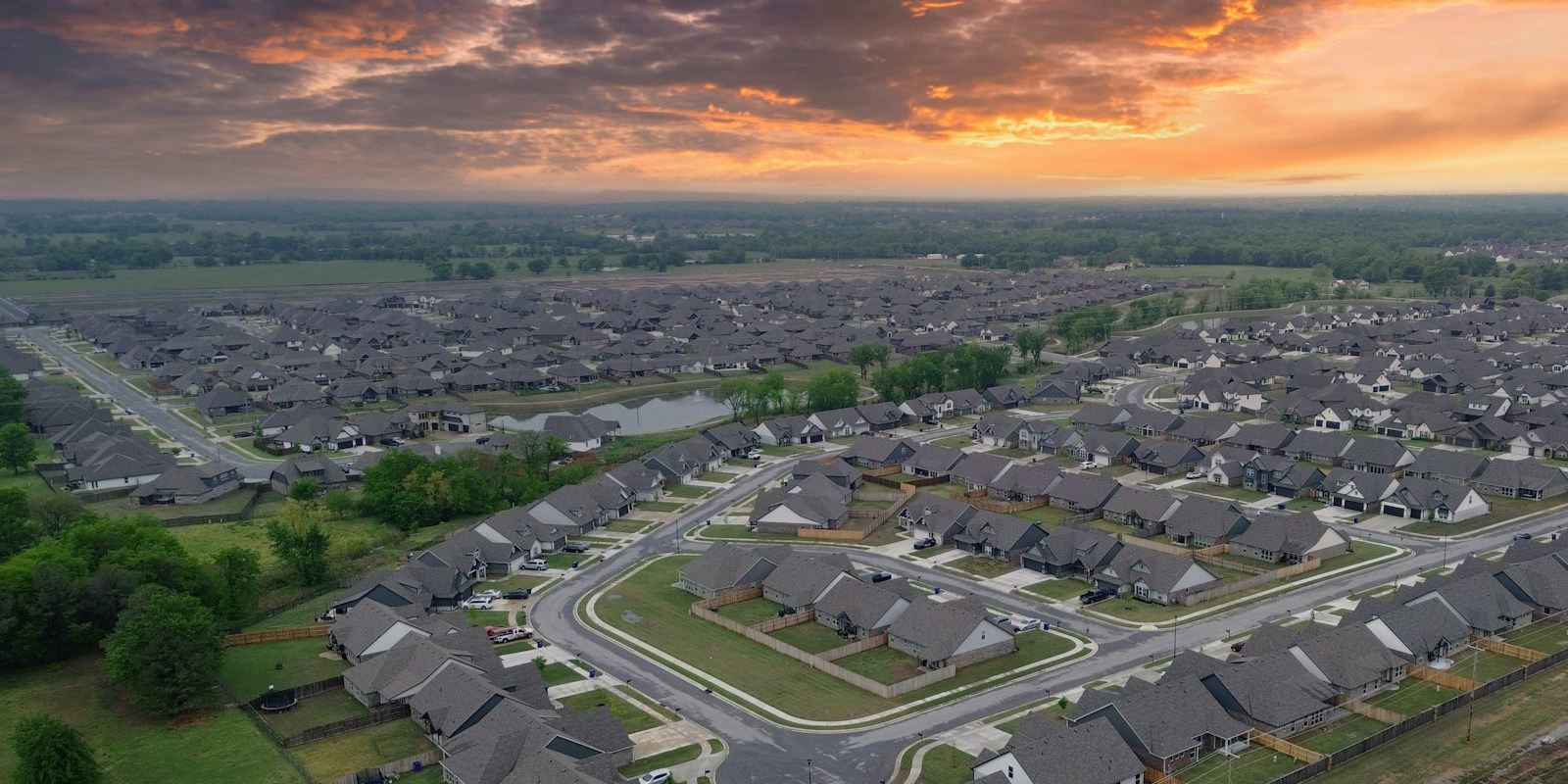 An aerial view of a suburban neighborhood in Broken Arrow, Oklahoma, featuring modern brick homes with dark roofs, winding residential streets, and a community pond under a vibrant sunset sky.